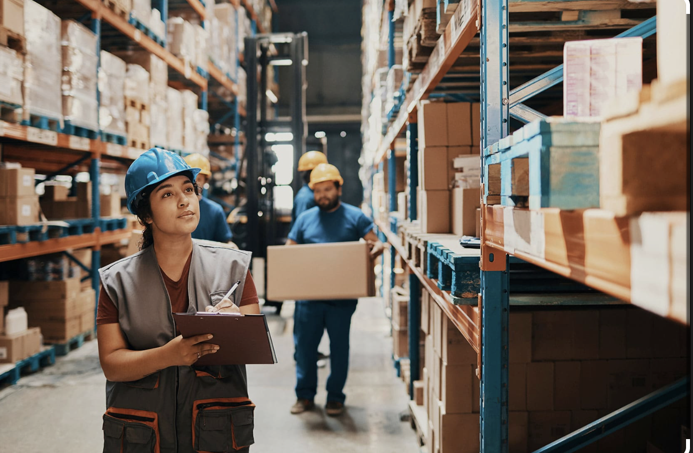 Lagerarbeiterin mit Schreibbrett erfasst Bestände im Hochregallager während Team im Hintergrund arbeitet – Personalvermittlung Lagerarbeiter