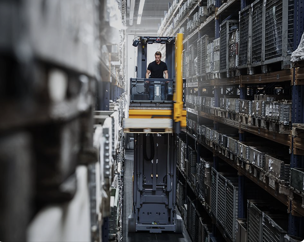 Staplerfahrer fährt mit hochgefahrener Kabine durch Hochregallager zwischen Regalgängen – Personalvermittlung Staplerfahrer