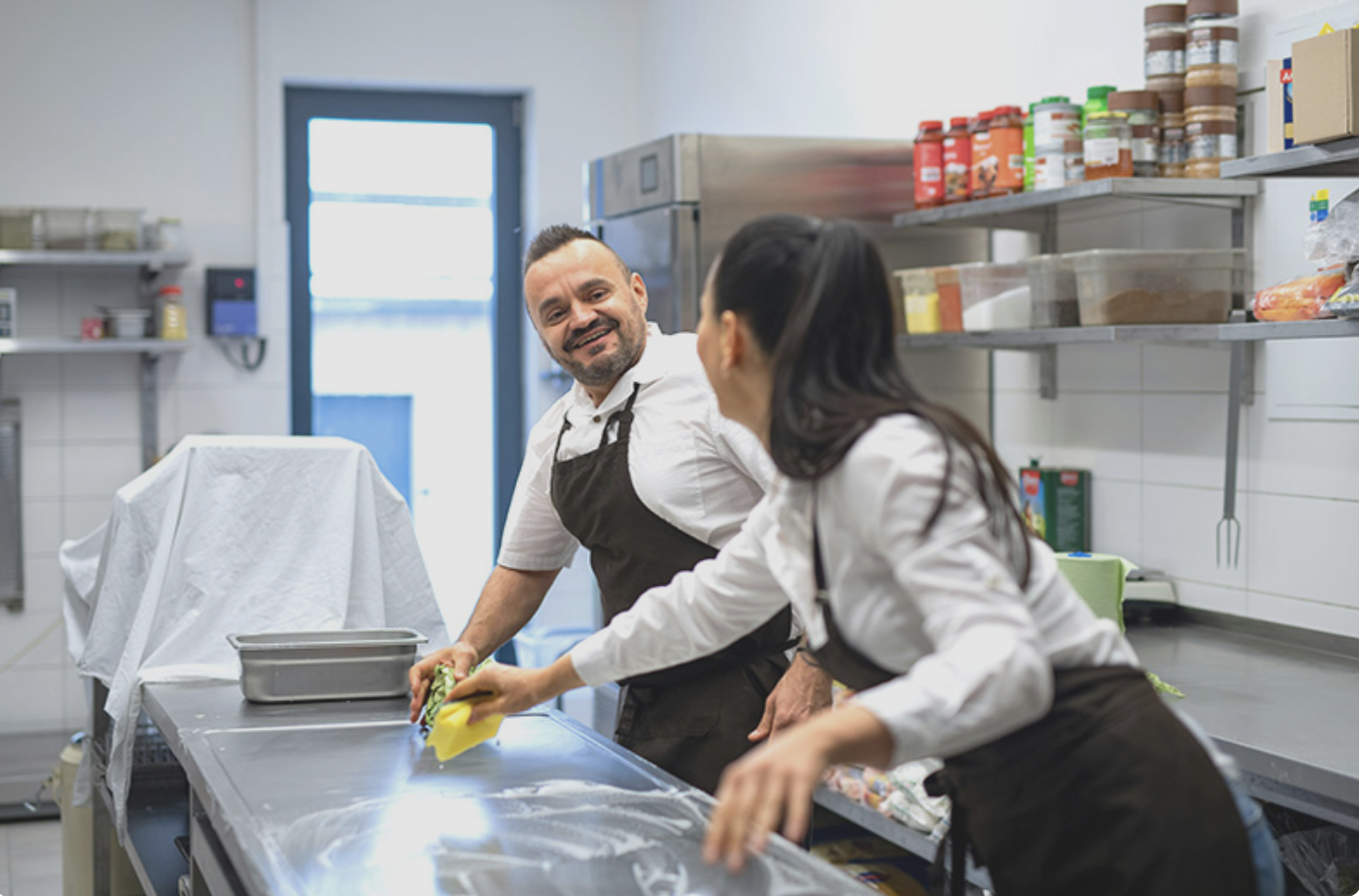 Mann und Frau reinigen gemeinsam eine große Arbeitsplatte in einer modernen Großküche